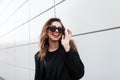 Pretty joyful young hipster woman in stylish sunglasses with a smile in a fashionable black coat posing outdoors