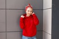 Pretty joyful young woman with a beautiful smile in a red sweater with a vintage bandana is standing near a gray wall