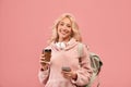 Pretty lady posing with backpack, holding coffee to go and cellphone, smiling at camera over pink studio background Royalty Free Stock Photo