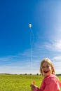 Pretty laughing little girl with blond hair on field in pink sweatshirt looks at colorful dragon in blue sky with soft clouds