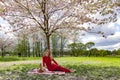 Pretty laughing woman in a long red dress sits under a blooming sakura tree in Victory Park