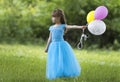 Pretty little blond long-haired girl in nice long blue evening dress holds colorful balloons standing in blooming field on blurred Royalty Free Stock Photo