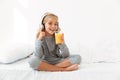 Pretty little blonde girl in gray pajamas holding glass of orange juice, while listening to music in bedroom Royalty Free Stock Photo