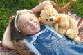 Pretty little child girl laying down with her teddy bear toy on blanket on green grass in summer smiling happily Royalty Free Stock Photo