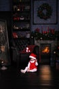 Pretty little child in santa cap near Cristmas tree. the baby girl sits on the floor near the fireplace and chairs alone as a smal