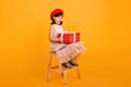 Pretty little girl in dress sitting on chair and holding big present box. Studio shot of french kid with birthday gift Royalty Free Stock Photo