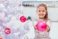 Pretty little girl is extending a pink bauble to the camera, standing near Christmas tree, smiling.