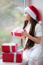 Pretty little girl in Santa hat dreaming by the window holding gift Royalty Free Stock Photo