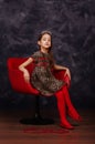 Pretty little girl wearing beautiful dress sitting in red armchair. She is wearing red masquerade carnival mask. Studio shot. Royalty Free Stock Photo