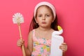 Pretty little girl wearing summer dress wants to halp her mommy to wash toilet, holding brush and detergent, looks at camera with