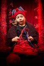 A pretty little girl who looks like a dwarf or elf is knitting with multicolored threads from large clubs on a black and red