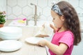 Pretty little girl wiping clear white plates with dry towel on kitchen set. Dry tableware after washing.