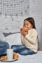 Pretty little girl 6 years old in a white sweater and jeans. Child in the room with a bed, eating croissant and drinking Royalty Free Stock Photo