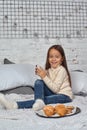 Pretty little girl 6 years old in a white sweater and jeans. Child in the room with a bed, eating croissant and drinking Royalty Free Stock Photo