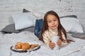 Pretty little girl 6 years old in a white sweater and jeans. Child in the room with a bed, eating croissant and drinking Royalty Free Stock Photo