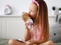 Pretty little kid girl in home clothing dress and headband sits cross legged playing with dolls kissing favorite at home Royalty Free Stock Photo