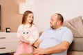 Pretty little lady with red hair standing near her father while he sitting on the sofa and girl holding in her hands Royalty Free Stock Photo