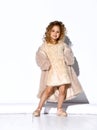 Pretty little princess with golden curly hair posing in pink fluffy dress and cape tilting her head and putting hands on hips, Royalty Free Stock Photo