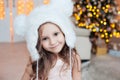 Pretty little smiling girl wearing white fur hat posing against Christmas tree indoors. Royalty Free Stock Photo