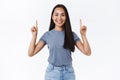 Pretty, lively smiling emotive brunette girl in casual blue t-shirt, pointing fingers up as fully pleased with product