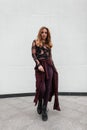 Pretty luxurious young woman with purple cape in a vintage patterned blouse in stylish pants in boots posing near a white wall