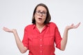 Pretty mature caucasian woman in red shirt and glasses shrugs shoulders with open palms
