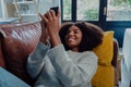 Pretty mixed race female smiling while scrolling on smartphone resting on leather sofa in trendy lounge.