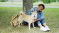 Pretty mixed race woman is fussing her pet dog resting on the grass in the park on warm summer day. Beautiful green lawn