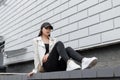 Pretty modern young woman in black stylish cap in a leather jacket in jeans in fashionable sneakers with a gold backpack resting Royalty Free Stock Photo