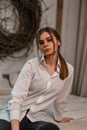 Pretty modern young woman in a fashionable white shirt in black vintage jeans is sitting on the bed in the room. Cute stylish girl Royalty Free Stock Photo