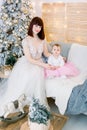 Pretty mother with her baby girl, wearing in fashionable elegant dresses, sitting on stylish gray sofa on the background Royalty Free Stock Photo