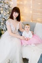 Pretty mother with her baby girl, wearing in fashionable elegant dresses, sitting on stylish gray sofa on the background Royalty Free Stock Photo