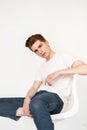 Pretty nice young man model with hairstyle in trendy white t-shirt in blue fashion jeans posing on chair near vintage wall indoors Royalty Free Stock Photo