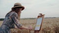 Pretty paintress in hat and dress painting landscape on canvas among wheat field