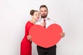 Pretty pleased couple standing together and hugging, man holding big heart, looking at camera. Royalty Free Stock Photo