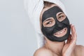 Pretty portrait of young beautiful woman applying facial cosmetic black clay organic mask on her face, wears white towel on hair. Royalty Free Stock Photo