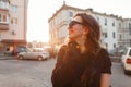 Pretty positive young hipster woman in stylish T-shirt in dark trendy sunglasses is standing and smiling outdoors near buildings Royalty Free Stock Photo