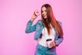 Pretty positive young woman in trendy blue jeans jacket with coffee with donut in sweet glaze posing near vintage pink wall. Royalty Free Stock Photo