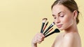 Pretty redhead teenage girl with freckles, looking down, holding diverse make up brushes. Model with light make-up.