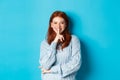 Pretty redhead teenager hushing and smiling, telling a secret, standing in sweater against blue background Royalty Free Stock Photo