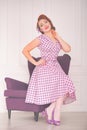 Pretty redheaded pin up woman wearing pink polka dot dress and posing with purple armchair on white background Royalty Free Stock Photo