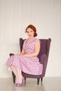 Pretty redheaded pin up woman wearing pink polka dot dress and posing with purple armchair on white background Royalty Free Stock Photo