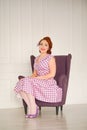 Pretty redheaded pin up woman wearing pink polka dot dress and posing with purple armchair on white background Royalty Free Stock Photo