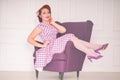 Pretty redheaded pin up woman wearing pink polka dot dress and posing with purple armchair on white background Royalty Free Stock Photo