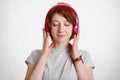 Pretty relaxed pleased ginger woman with short hair, closes eyes with pleasure while listens her favourite music with headphones,