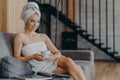 Pretty relaxed young woman looks thoughtfully into distance, holds cup of hot beverage, rests after showering, wrapped in bath