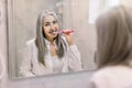 Pretty retired woman with long gray hair, wearing white shirt, brushing her teeth in the bathroom at home, standing in Royalty Free Stock Photo