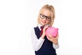 Pretty schoolgirl with blonde hair in school suit smiles and holds pink pig money box with money isolated on white Royalty Free Stock Photo