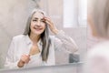 Pretty senior woman with long gray hair, wearing white shirt, looking at her face in the bathroom mirror, and applying Royalty Free Stock Photo