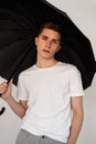 Pretty serious young nice man in trendy casual youth t-shirt stands with black vintage umbrella on white background in studio. Royalty Free Stock Photo
